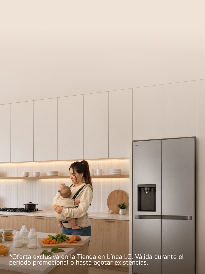 Una madre sostiene a su bebé en brazos en una cocina moderna, junto a un refrigerador LG, mientras prepara verduras frescas sobre la encimera.