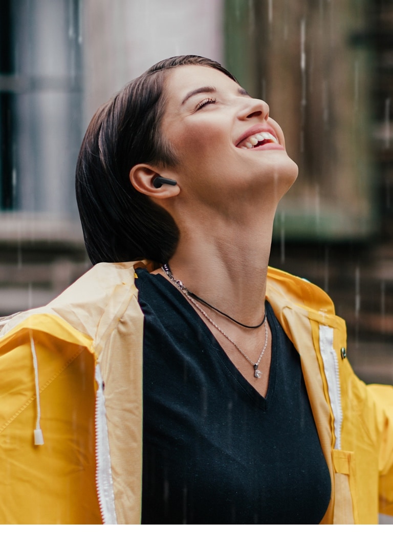 Une femme portant un imperméable de couleur vive utilise ses écouteurs tout en restant debout sous la pluie.
