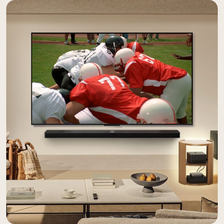 Salon z LG True Wireless TV na ścianie. Na ekranie mecz futbolu amerykańskiego. Konfiguracja telewizora zawiera również naścienny LG Soundbar. Prawie niewidoczne pozostałe urządzenia peryferyjne podłączone są do Zero Connect Box umieszczonego na bocznym stoliku. Cały salon jest schludny, uporządkowany i bez plątaniny kabli.