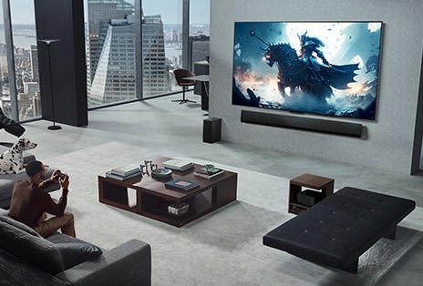 Salon z mężczyzną grającym na LG True Wireless TV i LG Soundbar zamontowanym na ścianie. Trzyma pada, ale konsoli nie widać. Nie widać kabli. Moduł Zero Connect Box jest schowany pod stolikiem, ledwo widoczny.
