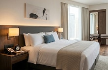Modern hotel bedroom interior featuring a neatly made bed, soft lighting, armchairs, and large windows with natural light.