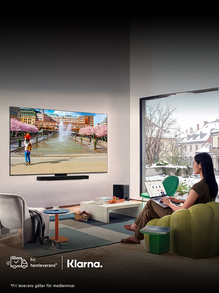 LG platt smart-TV monterad på väggen i ett vardagsrum, som visar en färgstark stadsscen, medan en person sitter i en grön soffa och tittar.