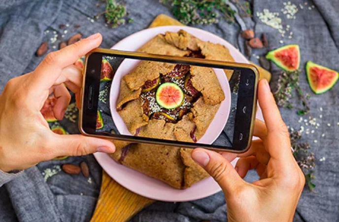 A pair of hands hold a smartphone over a fresh fig pastry while snapping a picture of the dish.