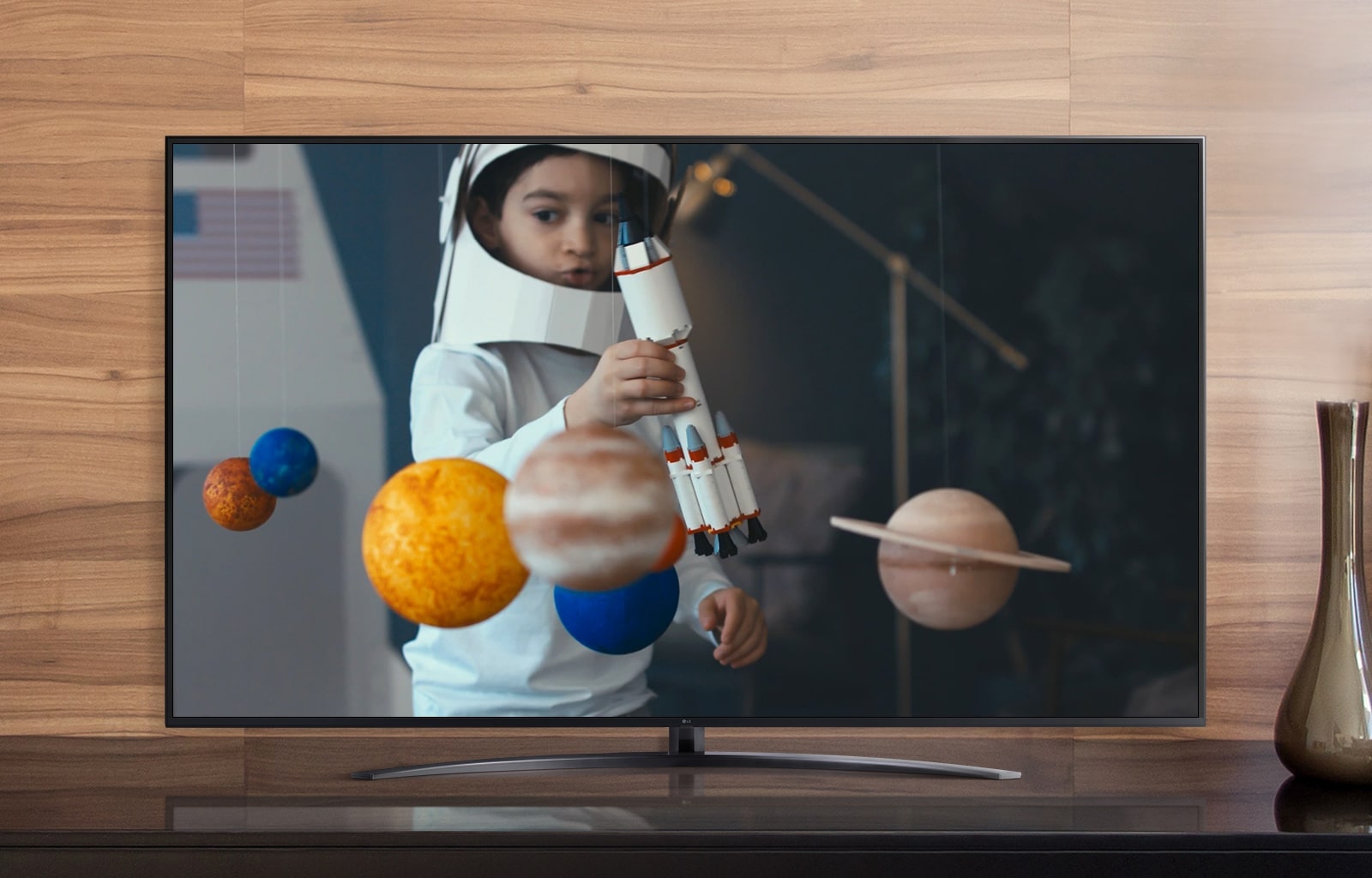 Un écran de télévision diffusant une vidéo d'un garçon vêtu d'un costume d'astronaute qu'il a fabriqué en jouant avec un vaisseau spatial dans sa chambre décorée de miniatures de planètes (jouer la vidéo)