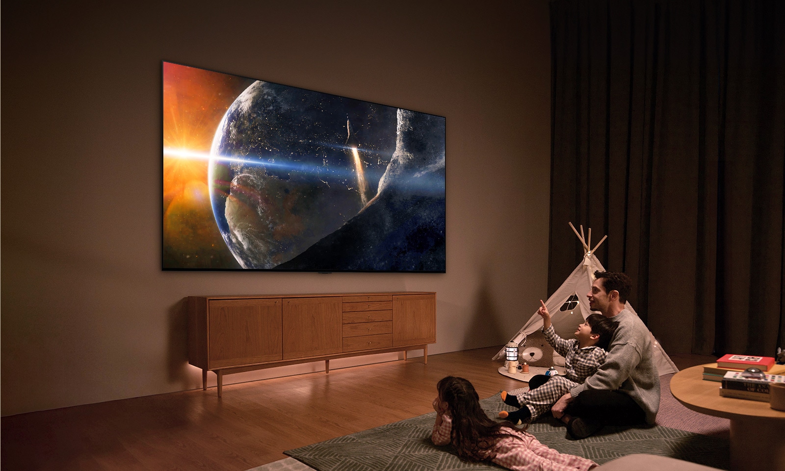 A family sat on the floor of a low-lit living room by a small table, looking up at an LG TV mounted on the wall showing the Earth from space.