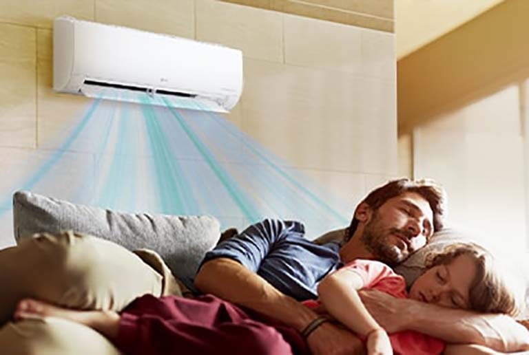 A father and daughter sleep on a couch beneath an air conditioner that is blowing air out over them.