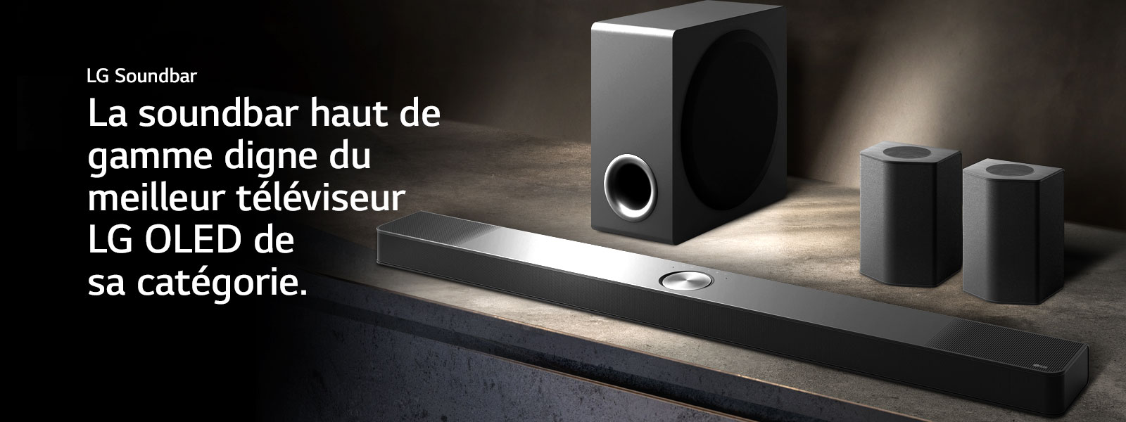 La LG Soundbar, les enceintes arrière et le caisson de basse installés en biais sur une étagère en bois marron dans une pièce obscure, avec une lumière qui n’éclaire que le système audio.