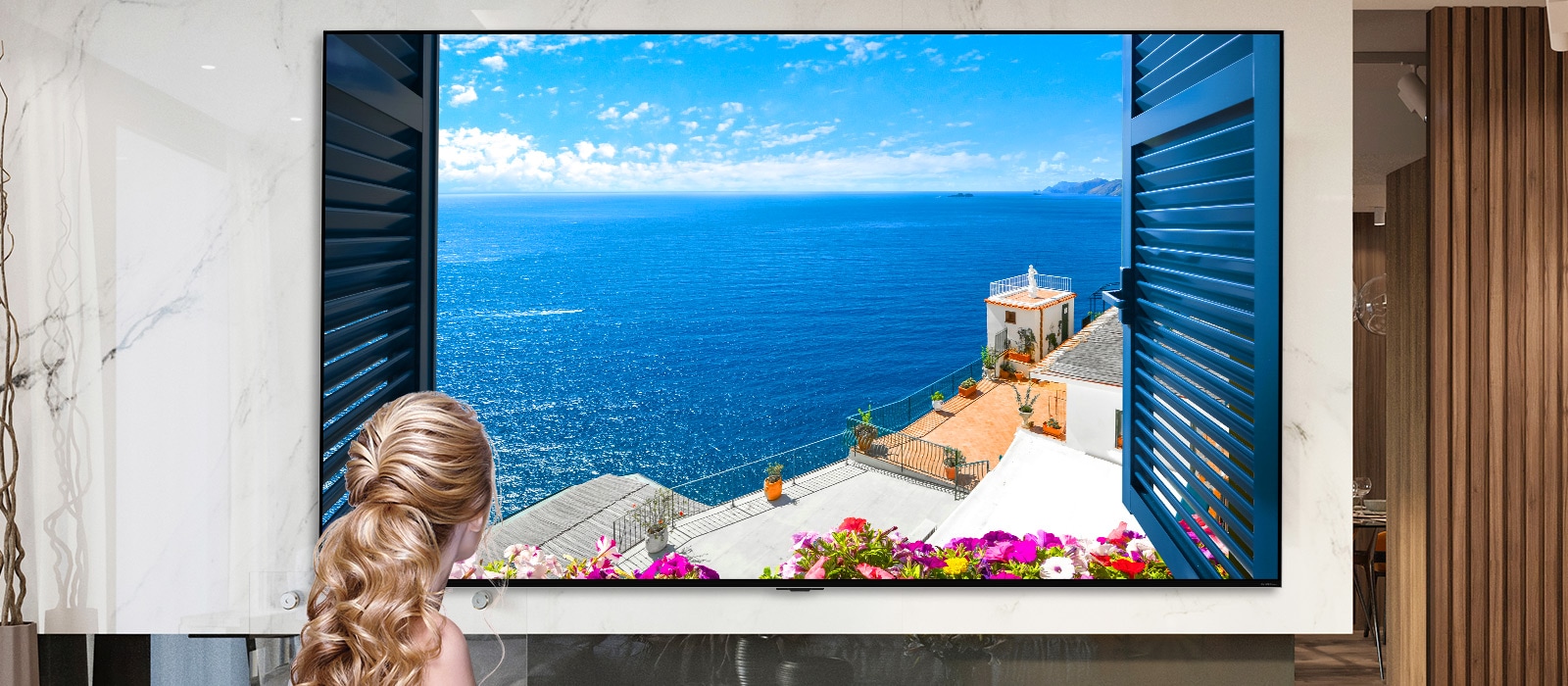 Vista trasera de una mujer mirando por la ventana un mar azul y edificaciones de color blanco. Al deslizarse en la página hacia abajo, la imagen se aleja para mostrar más de la habitación y revelar que la ventana es un televisor LG QNED Mini LED.