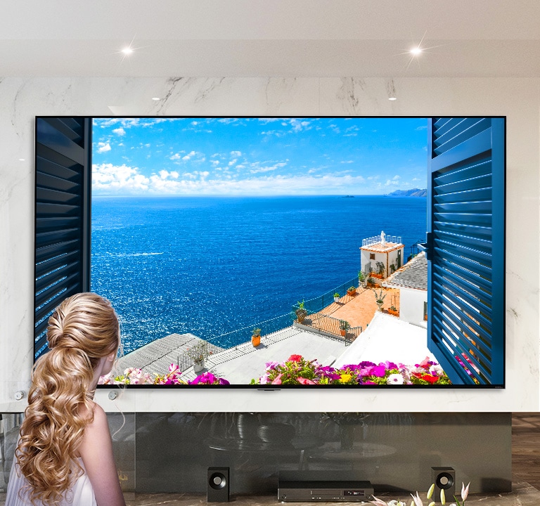 Vista trasera de una mujer mirando por la ventana un mar azul y edificaciones de color blanco. Al deslizarse en la página hacia abajo, la imagen se aleja para mostrar más de la habitación y revelar que la ventana es un televisor LG QNED Mini LED.