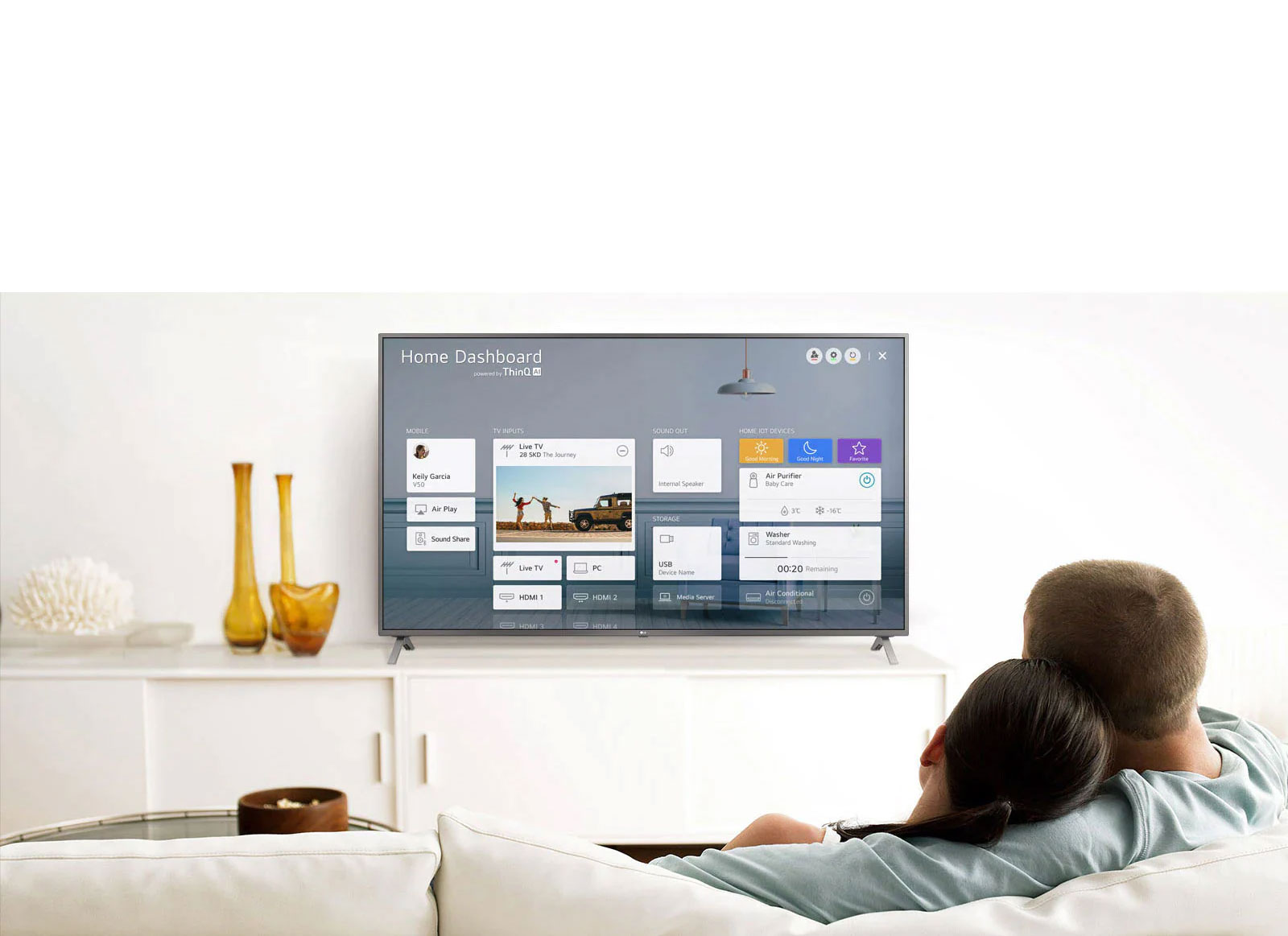 A men and women sitting on a sofa in the living room with the Home Dashboard on the TV screen.