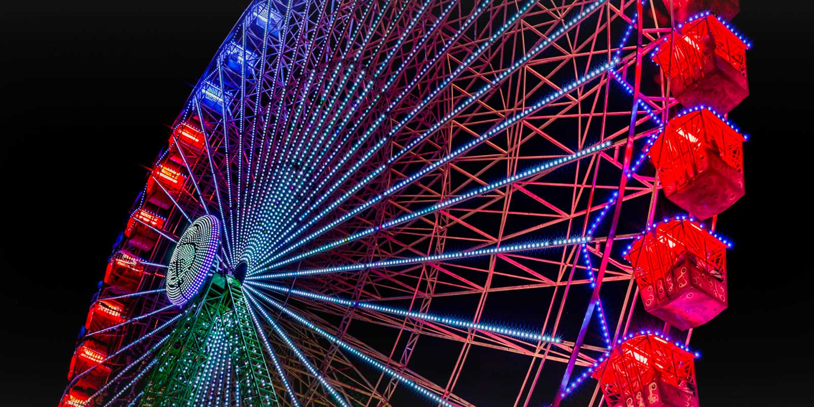 A brightly-lit, rainbow-colored ferris wheel against a black night sky, captured from a low angle.