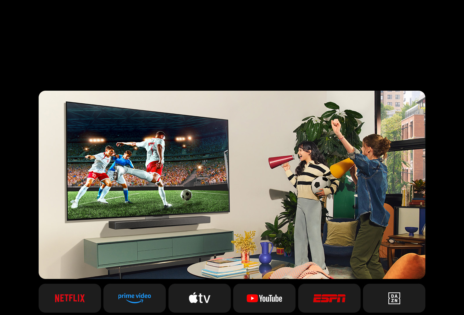 Two women watch a soccer game in a cozy living space. One woman holds a football and both cheer on their team with yellow and red loudspeakers. The following logos are shown below. Netflix, Amazon Prime Video, Apple TV, Youtube, ESPN, and DAZN.