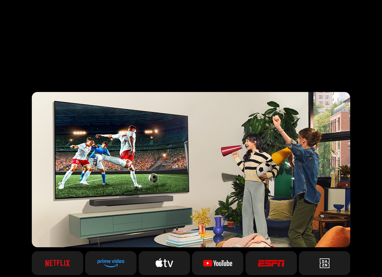 Deux femmes regardent un match de football dans un espace confortable. Une femme tient à ballon de football et toutes deux supportent leur équipe avec des haut-parleurs jaunes et rouges. Les logos suivants sont affichés en dessous. Netflix, Amazon Prime Video, Apple TV, Youtube, ESPN et DAZN.