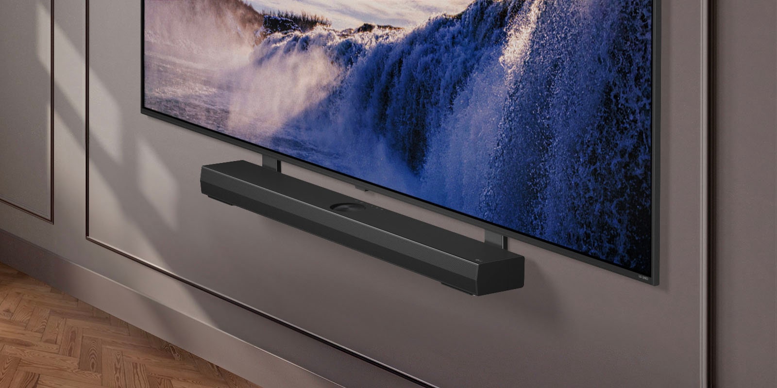 Une LG TV apparaît avec le support Synergy. Le support Synergy et la LG TV sont connectés. La caméra fait un zoom sur le support Synergy, révélant la barre de son, qui est placée sur le dessus du support Synergy, suivie de l’arrière-plan d’un espace de vie moderne.