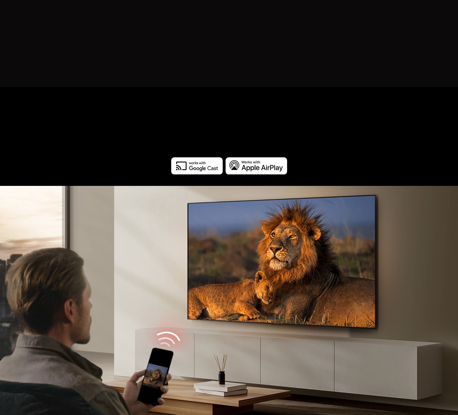 An LG TV mounted on a wall in a living room, displaying a lion and lion cub. A man sits in the foreground with a smartphone in his hand displaying the same image of lions. A graphic of three neon red curved bars is displayed just above the smartphone pointing toward the TV.