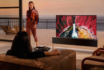 Eine Frau sitzt auf dem Sofa und schaut sich eine lebhafte Naturszene auf einem aufrollbaren OLED TV R an, während eine andere Frau daneben steht.