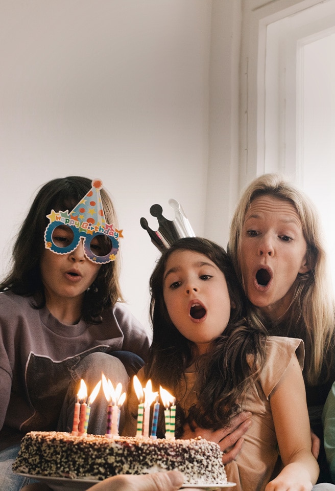 Image de deux femmes adultes et d’une jeune fille avec un chapeau d’anniversaire sur la tête et soufflant les bougies du gâteau.