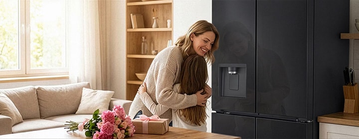 A mother and a child embracing next to a stainless-steel LG refrigerator, beside a bouquet of flowers and a gift on a table.