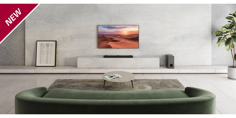 Ein Fernseher hängt an einer weißen Wandverkleidung in einem Wohnzimmer. Darunter steht eine LG Sound Bar auf einem TV-Regal. Auf dem Boden rechts vom Regal steht ein kabelloser Subwoofer. Auf Beistelltischen links und rechts hinter dem Sofa stehen zwei externe Lautsprecher. In der linken oberen Ecke steht „NEU“.