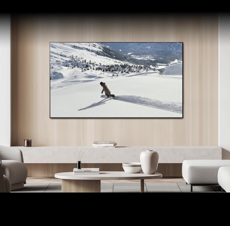 In einem elegant eingerichteten Wohnzimmer hängt ein Fernseher an einer holzvertäfelten Wand. Auf dem Bildschirm ist eine Snowboard fahrende Person zu sehen. Am unteren Bildschirmrand erscheint eine Sprechblase mit den Worten „Aktuelle Wiedergabe an Fernseher im Schlafzimmer senden“, woraufhin der Wohnzimmer-TV ausgeschaltet wird und die Sendung auf dem Fernseher im Schlafzimmer weiterläuft. Eine weitere Sprechblase mit den Worten „Aktuelle Wiedergabe an Fernseher im Wohnzimmer senden“ wird eingeblendet, woraufhin die Sendung wieder auf dem Wohnzimmer-Fernseher wiedergegeben und der Schlafzimmer-TV ausgeschaltet wird.