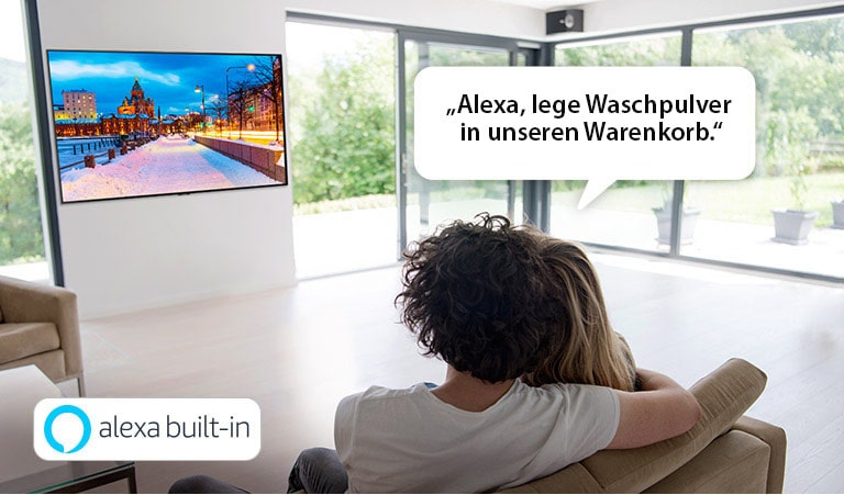 Ein Paar sitzt auf einer Couch und bittet Amazon Alexa, Waschpulver in seinen Warenkorb zu legen