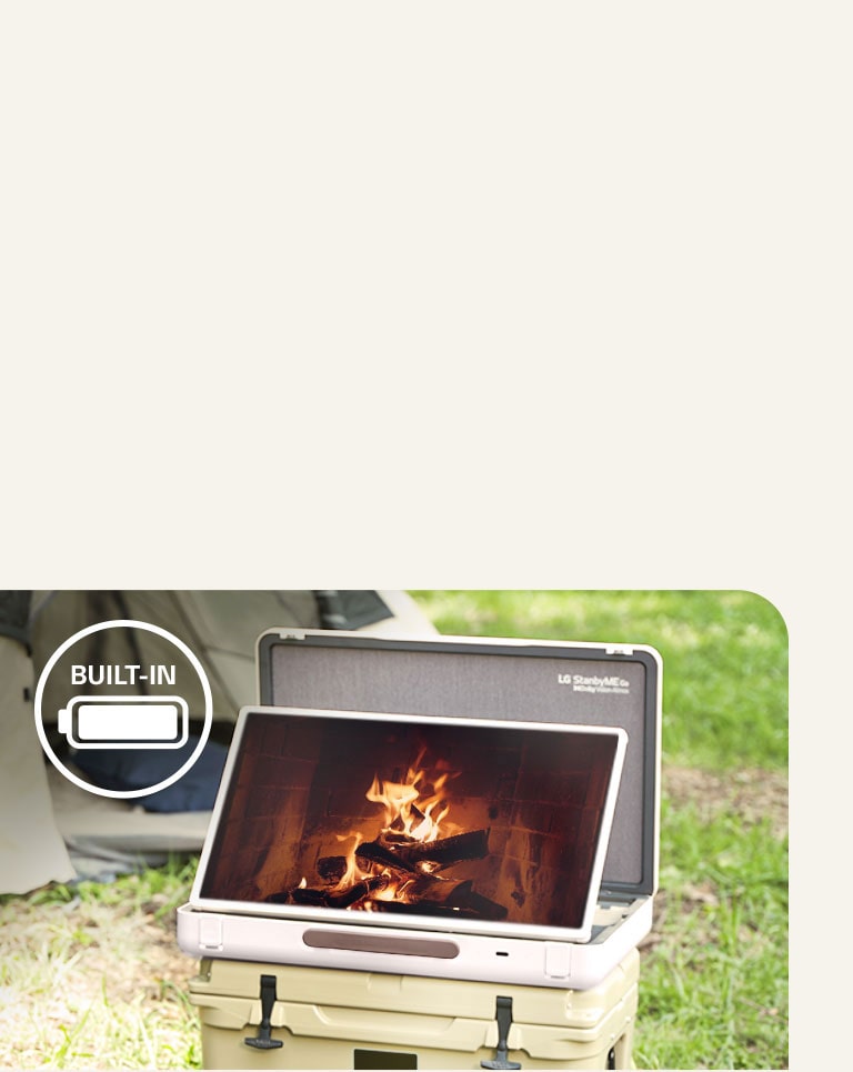 The LG StanbyME Go is placed in front of a tent, and the screen displays one of the relaxing themes--a fireplace. On the left top corner, built-in battery icon is shown.