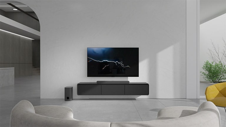 The sound bar is placed on the gray cabinet in the center and the connected TV is hung on the white wall right above, with lightnening image on it. White and red colored long sofa is placed facing the TV and the sun light is coming from the right side where a green plant is placed.