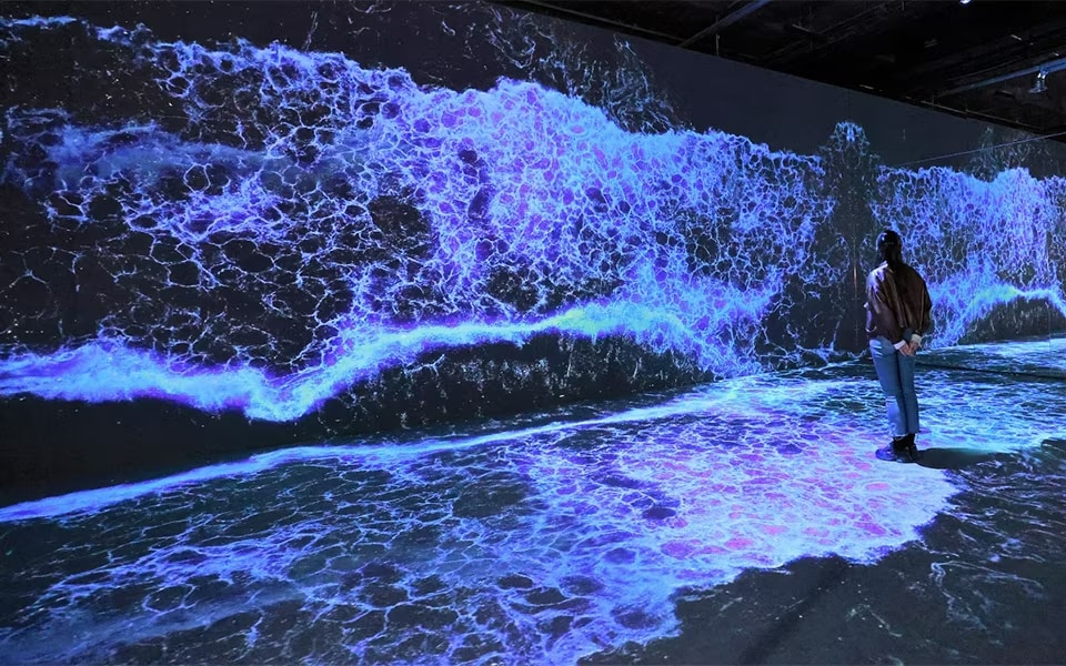 Person standing in front of a large, immersive digital display of crashing waves, showcasing vibrant blue and purple colours. The scene is projected onto a wall and floor, creating a dynamic and interactive visual experience.