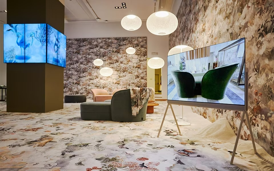 A stylish, modern room featuring floral-themed decor with patterned walls and carpet. Multiple LG OLED TVs display vivid images, including a close-up of green chairs. The room includes contemporary furniture, such as a pink chair and a green sofa, under soft, spherical hanging lights.