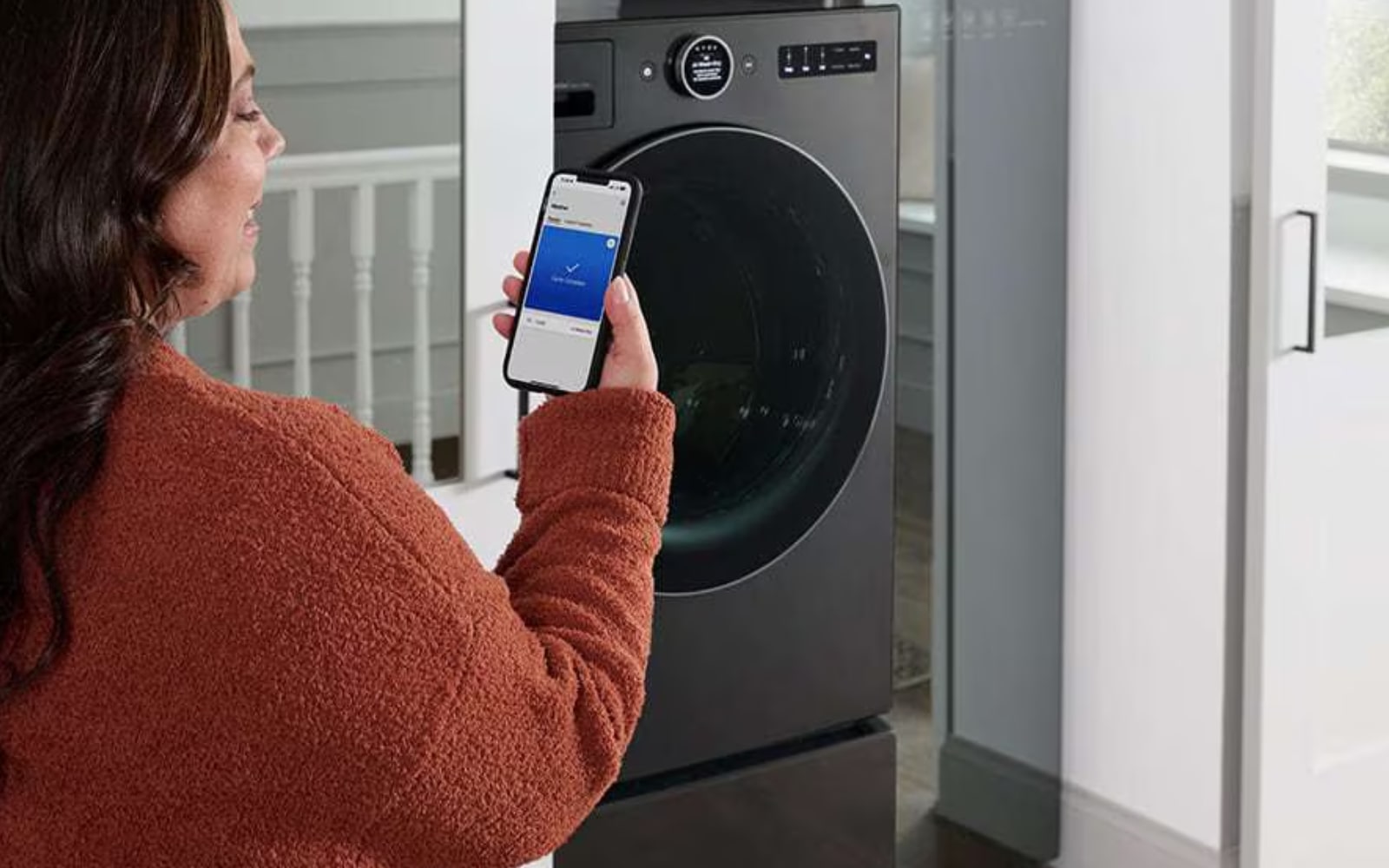 Person using the LG ThinQ™ app on their smartphone to control or monitor an LG washing machine. The person appears to be selecting a washing program remotely, highlighting the smart connectivity and convenience features of the appliance.