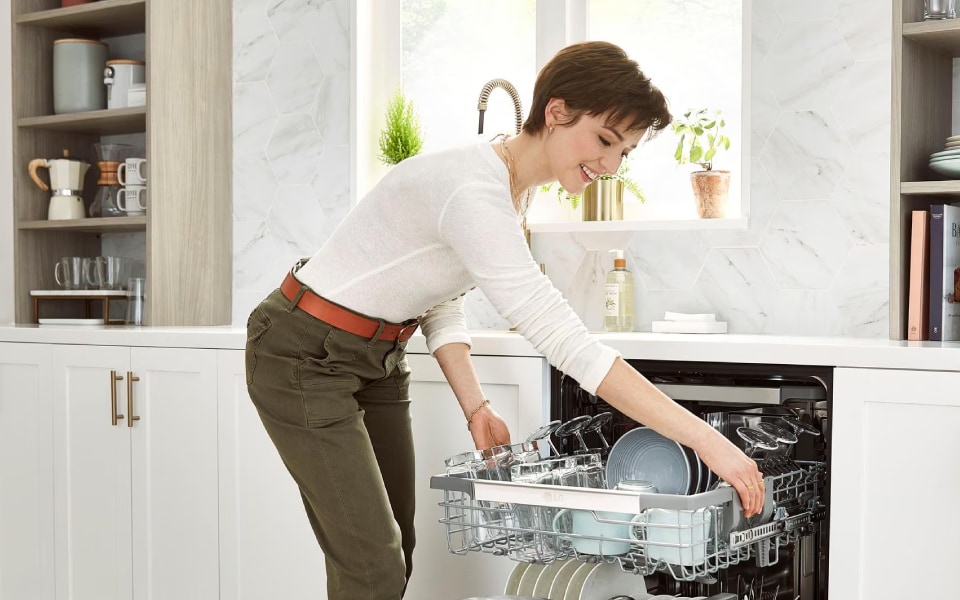 A woman is loading a dishwasher in a modern kitchen with white cabinets and a patterned rug on the floor. The dishwasher is fully open, and she is placing a dish on the top rack. 