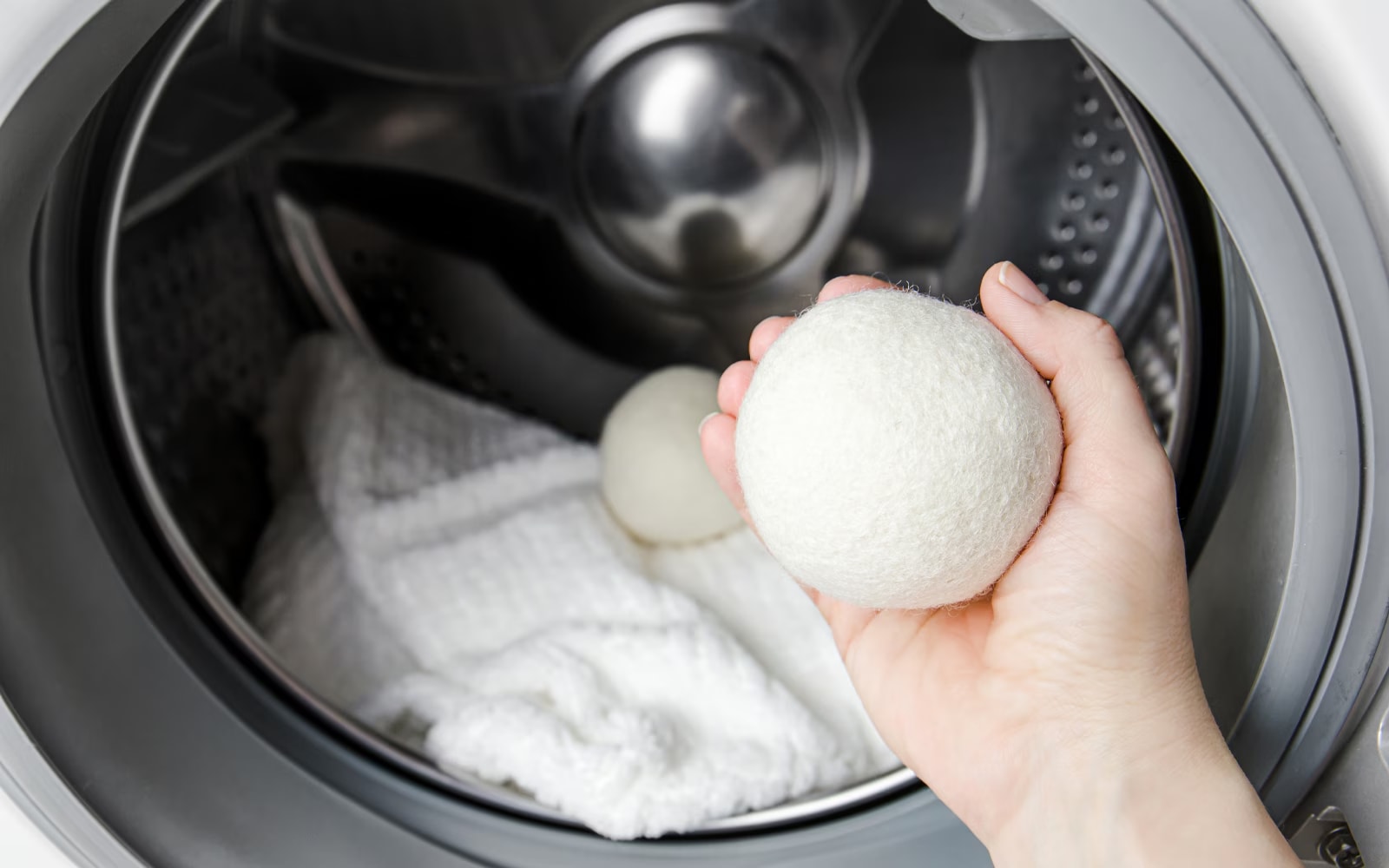 The inside of an LG Heat Pump dryer with a person holding a wool dryer ball in their hand. Inside the drum, there is a white towel along with another dryer ball.