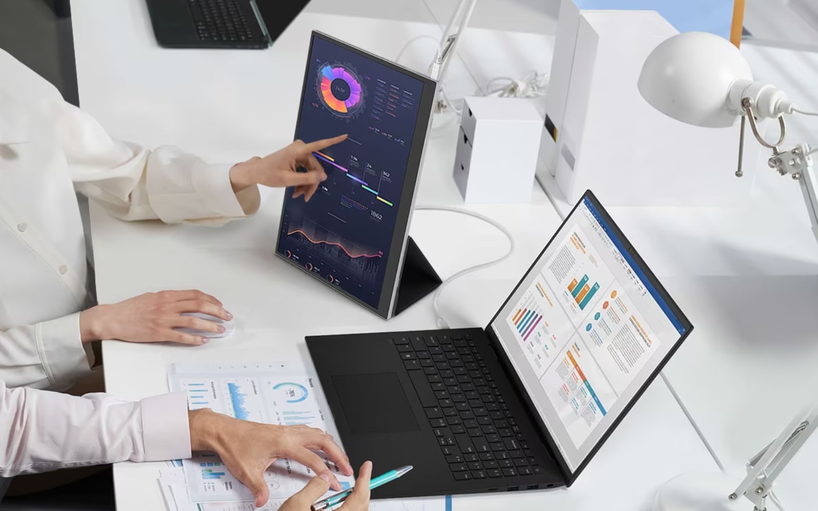 Two people work at a desk with a LG gram Laptop and +view Portable Monitor displaying charts, one points while the other uses a pen and mouse. Portable Monitor is available separately.  