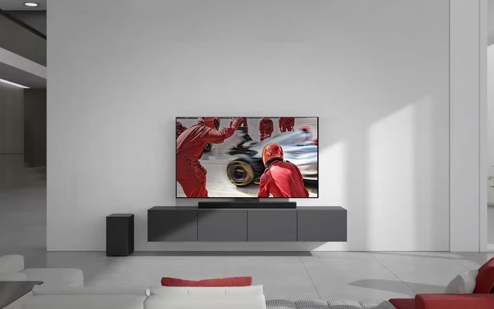 Modern living room with a wall-mounted flat-screen TV displaying a racing scene. Below the TV is a sleek soundbar on a floating console. The room features minimalist decor with a white sofa, a red cushion, and a small speaker on the floor.