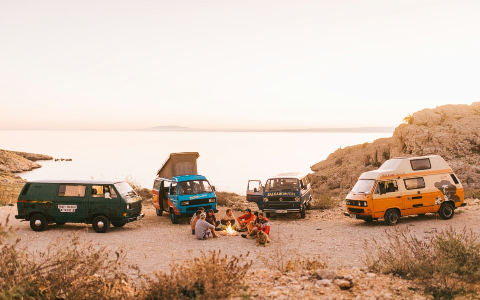 Group of people relaxing by a campfire near three colourful camper vans parked by a seaside at sunset.