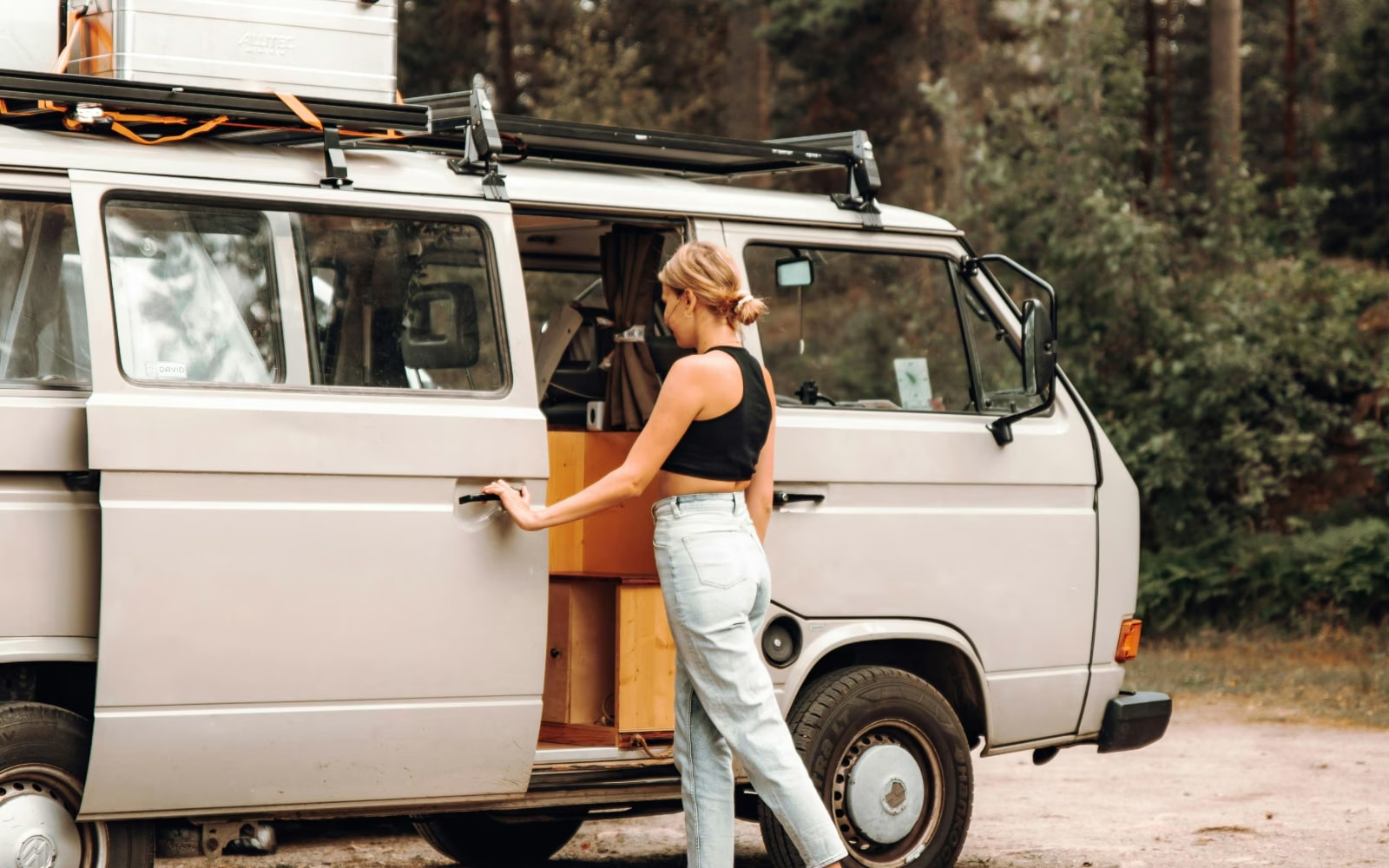 A woman opening the sliding door of a vintage camper van parked in a forest.