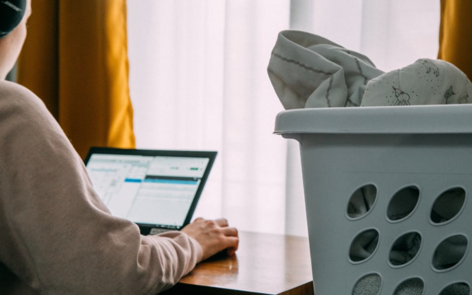 Person using a laptop next to a window with yellow curtains, while a laundry basket filled with clothes is in the foreground.