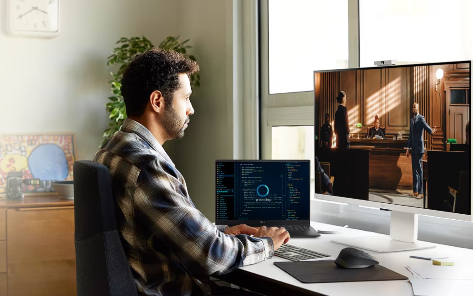 person working at a desk with an LG monitor and a laptop. The monitor displays a scene from a video or movie, while the laptop shows coding or data. The workspace is bright and organised, with plants, artwork, and a window providing natural light.