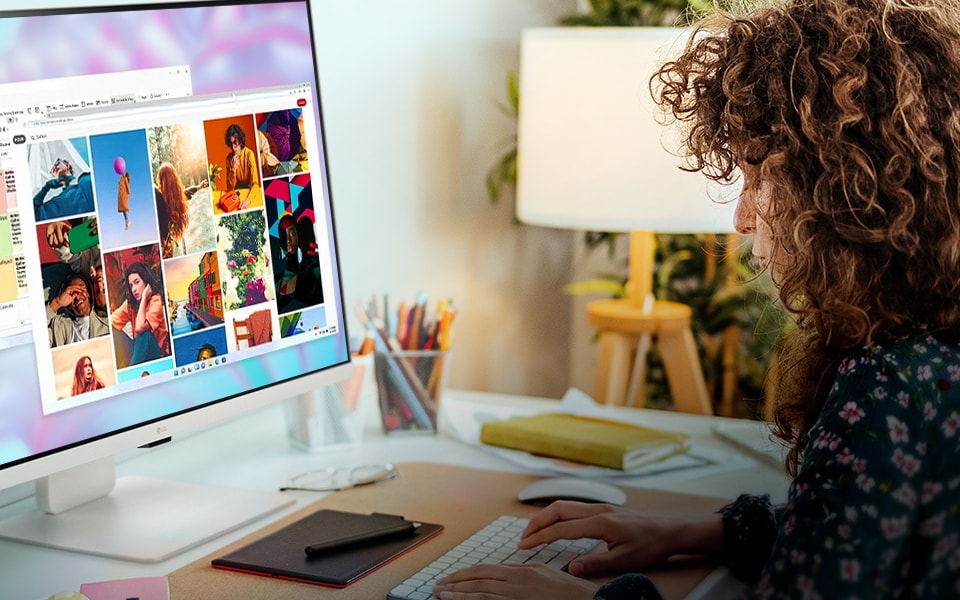  person working at an LG smart monitor, displaying images and documents. The desk setup includes a plant, lamp, and notebooks in a cosy home office environment.