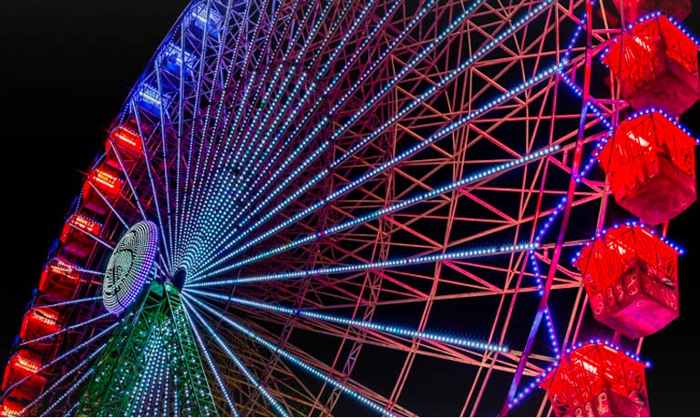 A brightly-lit, rainbow-colored ferris wheel against a black night sky, captured from a low angle.