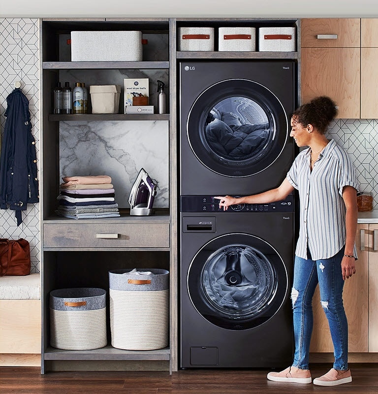 A black LG Wash Tower is installed in a laundry room and a woman stands to one side leaning in to press a button on the machine.