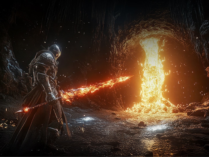 A heavily armored warrior stands in a dark, rocky cavern wielding a glowing, fiery sword. In front of the warrior, a massive torrent of molten lava pours down from the cave ceiling, casting a bright, intense orange light across the scene. The contrast between the darkness of the cave and the blazing fire emphasizes the game's dramatic and cinematic visual style.