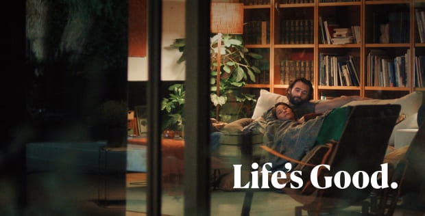 A man is sitting on a sofa in front of a bookshelf. A woman is leaning against the man's arm. The text Life's Good is written beneath the image.