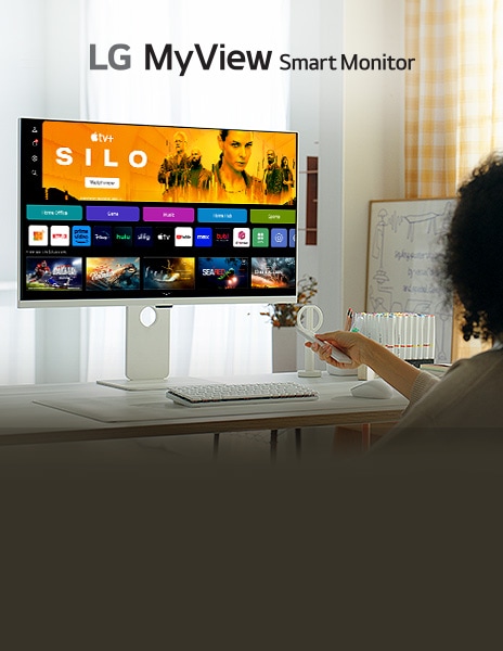 A woman sitting in front of a smart monitor placed on the table, pressing buttons on the remote control.