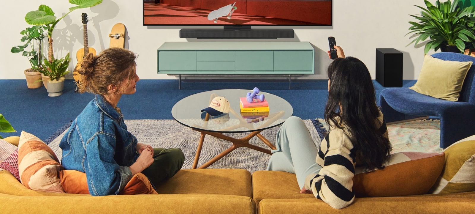 Two women sitting on a sofa in front of a wall-mounted LG TV in a colorful living room, and one woman points a remote at the TV screen. 	