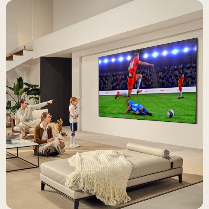 Uma família está na sala de estar. Eles têm uma LG TV montada na parede com uma tela ultra grande que é quase tão grande quanto a parede inteira. Eles estão assistindo a um emocionante jogo de futebol na tela grande e imersiva.