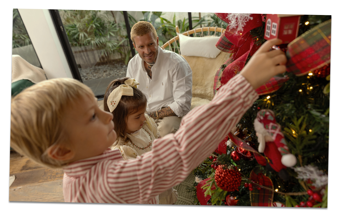 Um pai e as crianças estão se divertindo decorando uma árvore de Natal. O menino segura um enfeite para colocar na árvore.