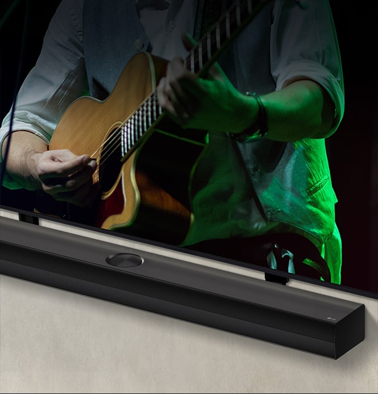 LG Soundbar contra um fundo preto revela seu design começando no canto esquerdo e depois se expande para mostrar toda a barra de som. Uma TV LG QNED aparece com Synergy Bracket. O Soundbar fica em cima do suporte Synergy, pressionado contra a parede com a tela inferior da TV visível, exibindo um homem tocando violão.