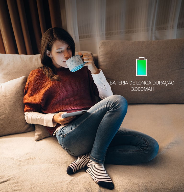 Mulher sentada em um sofá, tomando café e olhando o seu smartphone