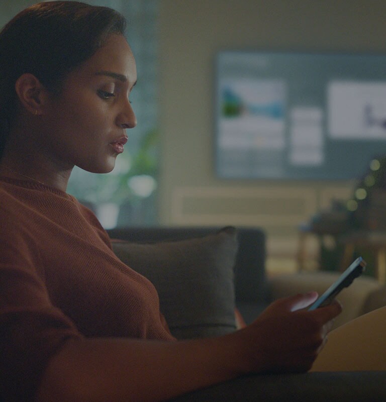 ] Imagem mostra uma mulher olhando para seu celular sentada no sofá da sala, enquanto a TV mostra o Homeboard ao fundo.